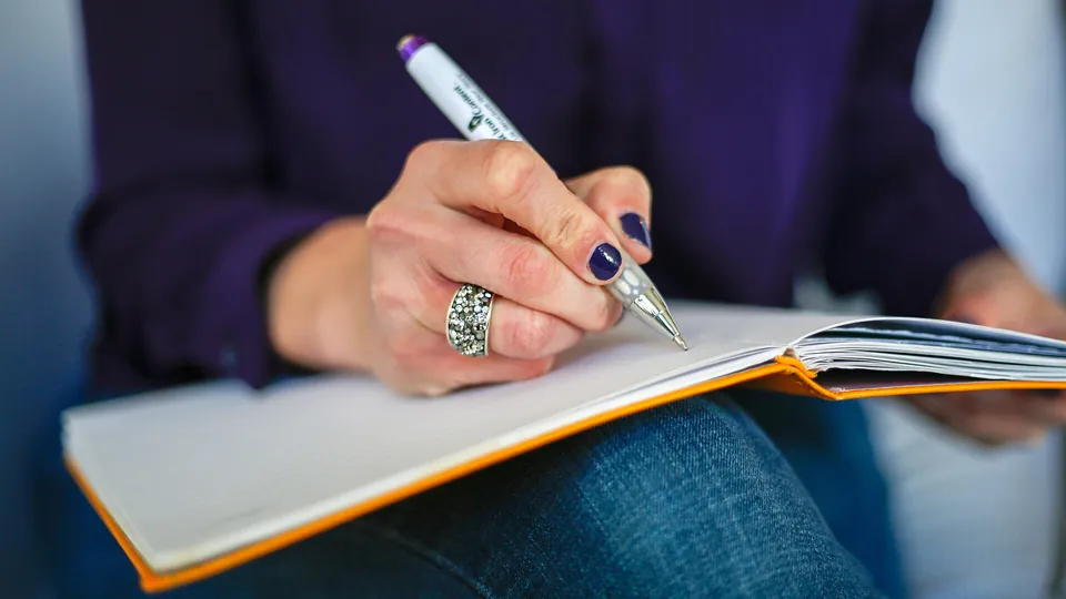 Eine Hand schreibt mit einem Stift in ein offenes Notizbuch, daneben liegt ein weiterer Stift.